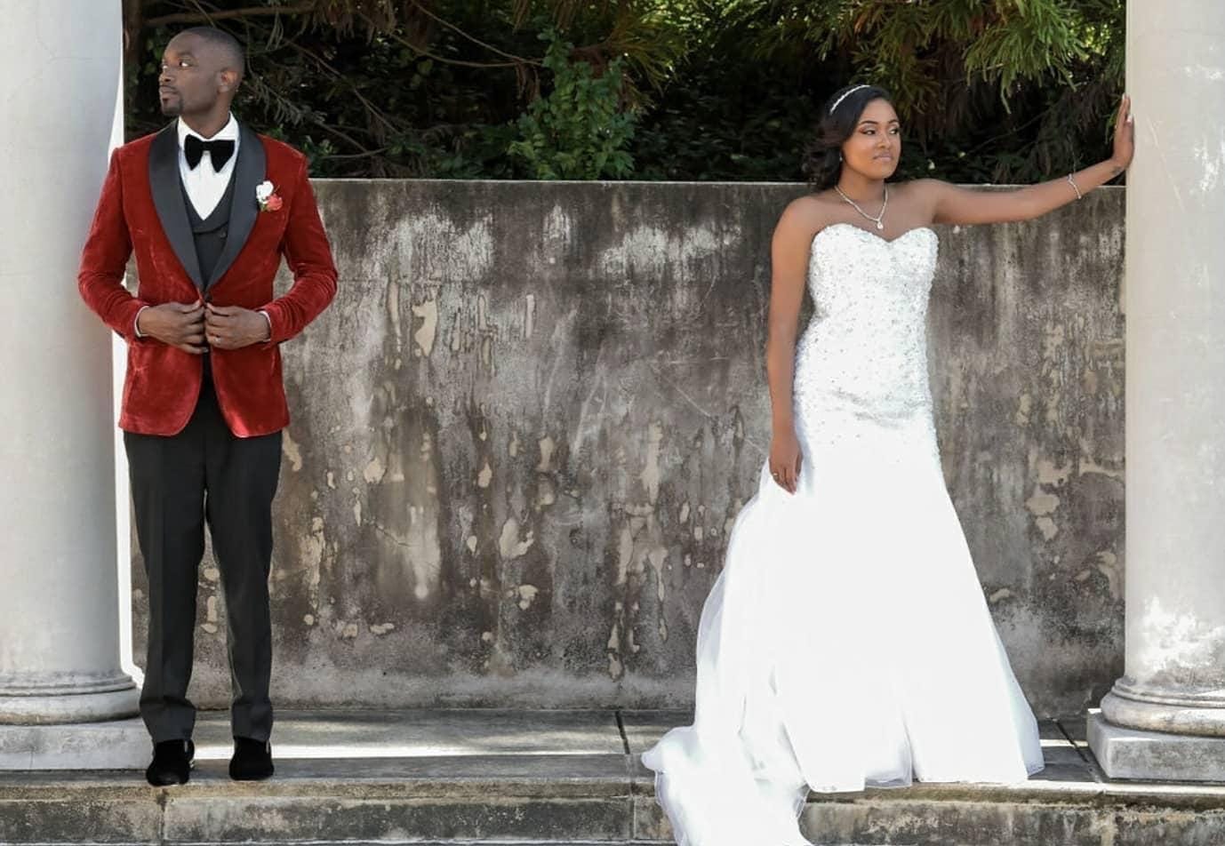 Groom in red velvet tuxedo with bride beside stone columns